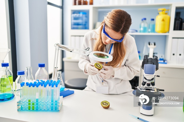 Young blonde woman wearing scientist uniform examining kiwi using magnifying glass at laboratory