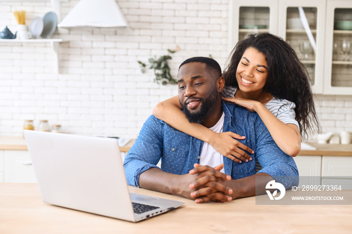 Biracial woman hugging man from behind, mixed ethnicity couple relax in new apartment at home browsing internet on laptop, show love everyday in the simplest of ways, watching video, looking at screen