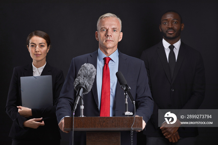 Waist up portrait of mature man giving speech standing at podium with two assistants in background