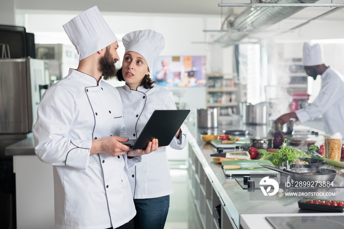 Food industry workers standing in restaurant professional kitchen while using computer to decide dinner service dish. Chefs looking for gourmet cuisine recipes on computer. Cooking concept