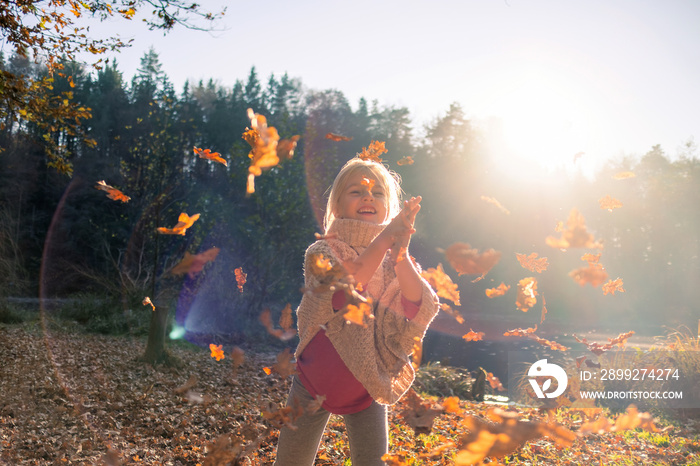 Smiling girl child throwing dry leaves in the air at beautiful autumn sunset.