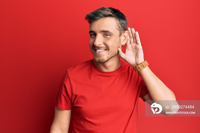 Handsome caucasian man wearing casual red tshirt smiling with hand over ear listening an hearing to rumor or gossip. deafness concept.