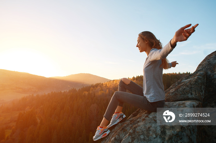 young woman sits on edge of cliff against background of sunrise