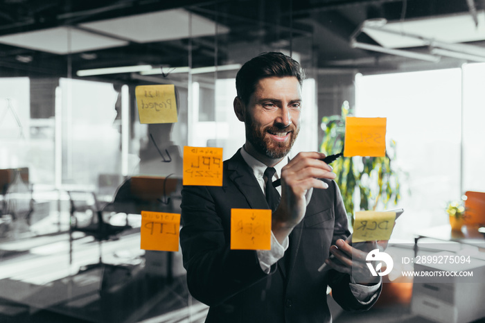 Successful businessman rewrites a task on a glass board with colored stickers, from phone and tablet