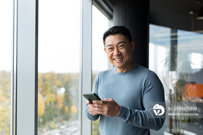 Happy and confident Asian businessman looking at camera and smiling, in modern office workplace, man holding phone