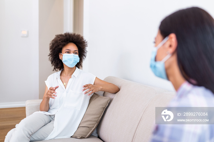 Two female best friends sitting in social distance wearing face mask and talking on the sofa, preventing covid 19 coronavirus infection spread.