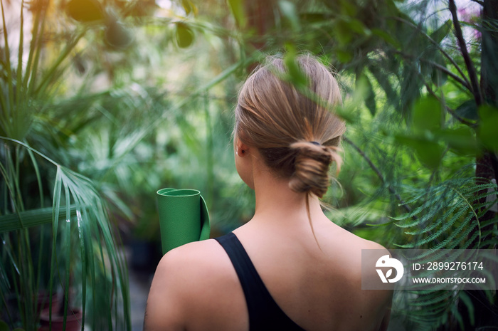 Portrait of cute young woman holding a rolled yoga mat in jungle. Sunny day. Back view