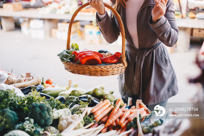 Woman holding basket full with vegetables and standing by stand at farmers’ market.