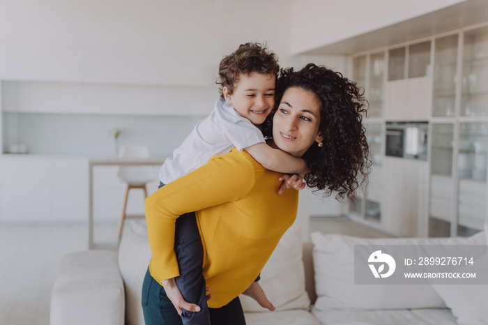 Young Spanish curly woman having fun with son, rolls him on back smiling wide. Babysitter in yellow sweater and green pants entertains little cheerful child at home. Elder sister plays with brother.