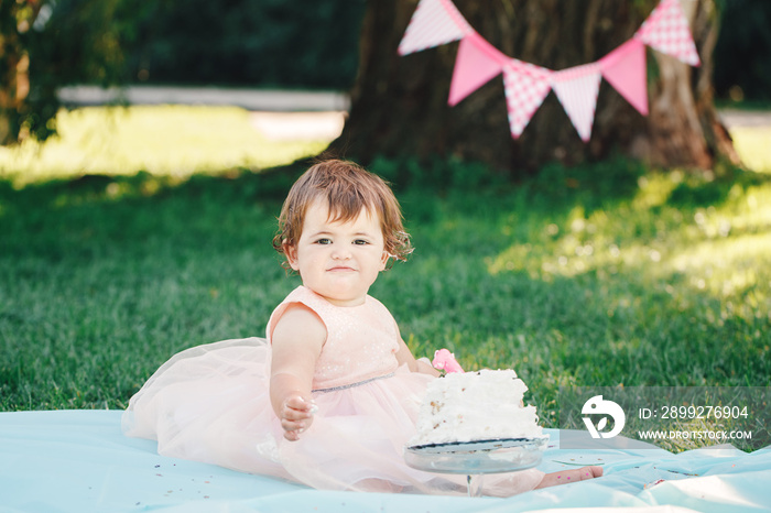 Portrait of cute adorable Caucasian baby girl with dark brown eyes in pink tutu dress celebrating her first birthday with gourmet cake looking in camera outside in park, cake smash