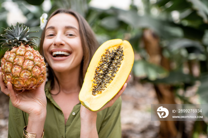 Portrait of a young smiling woman with sliced papaya and ananas outdoors on the tropical plantation. Concept of vegetarianism, healthy eating of fresh fruits and wellbeing