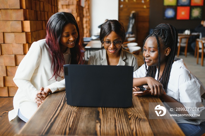 Three African American girls (students) sitting at the table in cafe studying up for test or making homework together, they are using laptop and digital tablet.