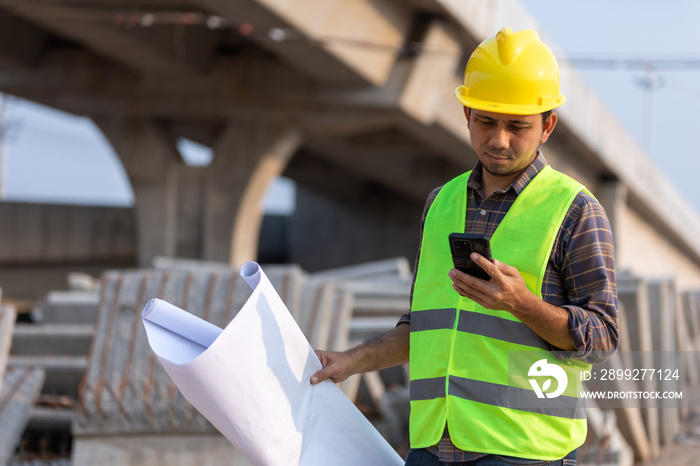 Asian construction workers use smartphone and hold a plan blueprint at the construction site