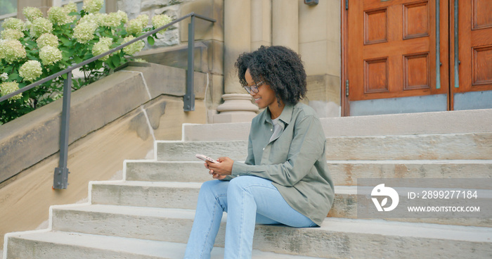 Afro American young student woman seats on the stairs and chatting in messenger, outdoors