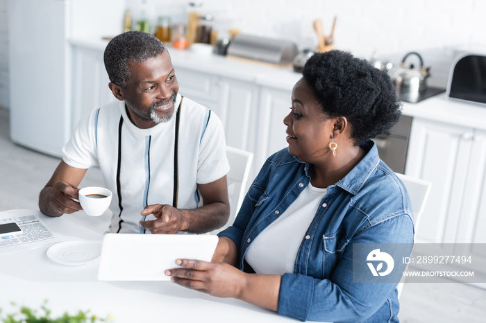 cheerful african american woman showing digital tablet to smiling husband with cup of coffee.