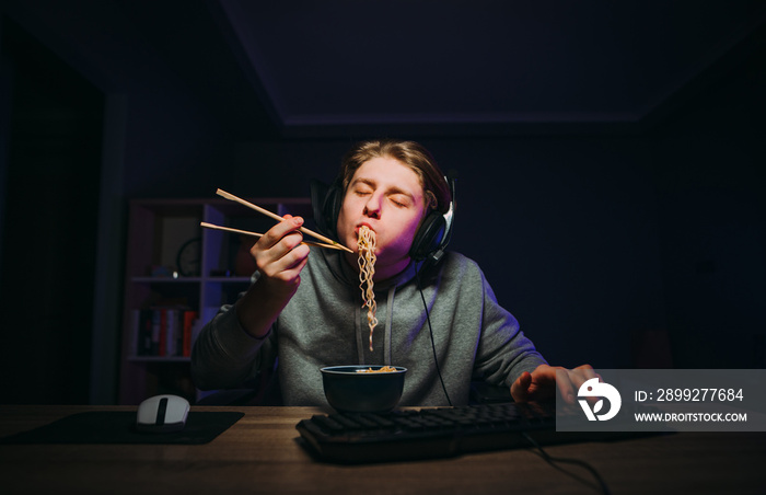 Hungry gamer guy in a headset sits at the computer at night and eats Chinese chopsticks with closed choi in a dark room