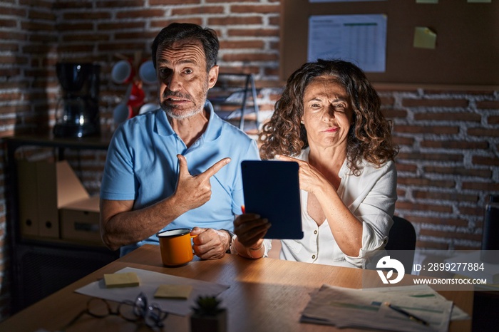 Middle age hispanic couple using touchpad sitting on the table at night pointing aside worried and nervous with forefinger, concerned and surprised expression