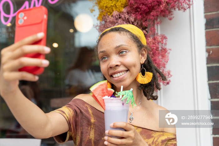 Young woman taking selfie while drinking smoothie in front of cafe