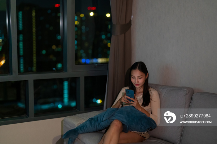 Asian woman sitting on couch in living room with using mobile phone with internet for social media or online shopping at night. Beautiful girl enjoy city life indoor lifestyle with technology at home