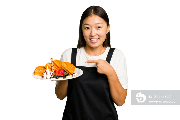 Young asian waitress woman holding waffles isolated smiling and pointing aside, showing something at blank space.