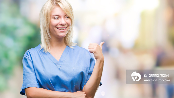 Young beautiful blonde nurse doctor woman over isolated background smiling with happy face looking and pointing to the side with thumb up.