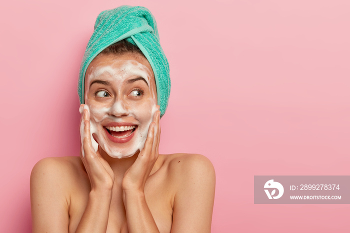 Headshot of lovely smiling female model touches cheeks, looks aside, washes face with soap bubble, has naked body, wears turquoise soft towel on head, poses over rosy wall, copy space for promotion