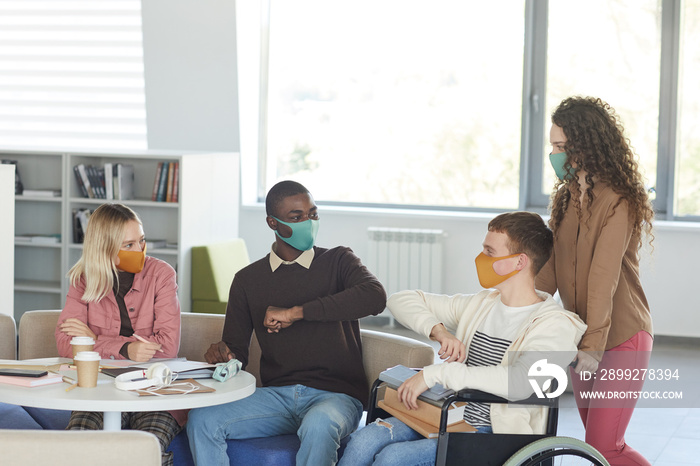 Side view at multi-ethnic group of students wearing masks while studying in college library with young man using wheelchair in foreground, copy space