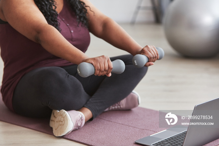 Cropped shot of curvy African American woman working out at home with dumbbells while sitting on yoga mat and watching online training videos, copy space