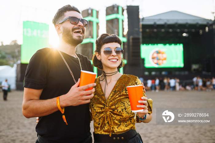 Stylish couple enjoying a performance at a festival together. Young people drinking beer and having fun at music festival at the beach. Youth, party, vacation concept.