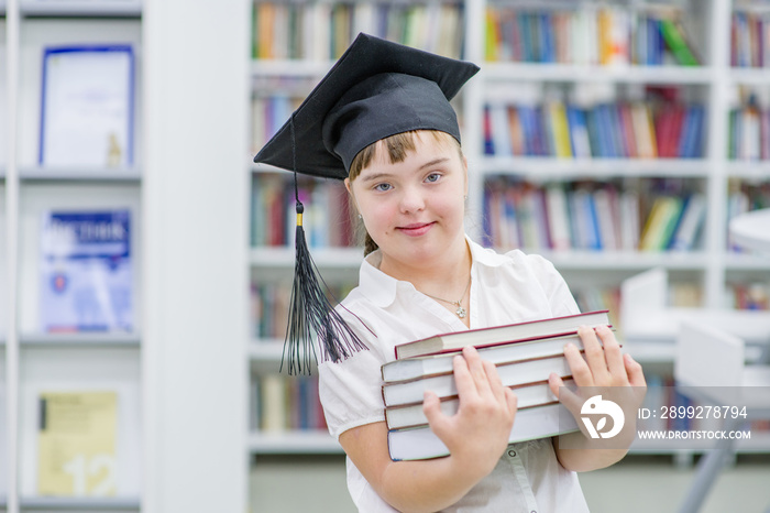 Happy young girl with Down Syndrome wearing a graduation cap holds books in her hands at a library. Empty space for text