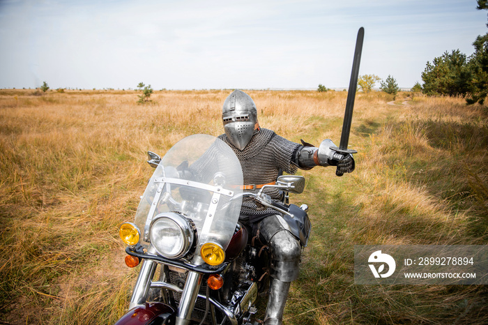 A medieval knight in chainmail and a helmet with a sword in his hands sits on a motorcycle.