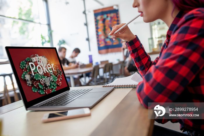 Side view of a young pink hair woman keyboarding on laptop computer with poker online on a screen while sitting in cafe.