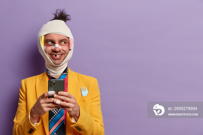 Positive victim of savage attack has hematoma, missing teeth and broken nose, holds smartphone in hands, spends rehabilitation period at home, wears yellow formal suit, isolated on purple wall