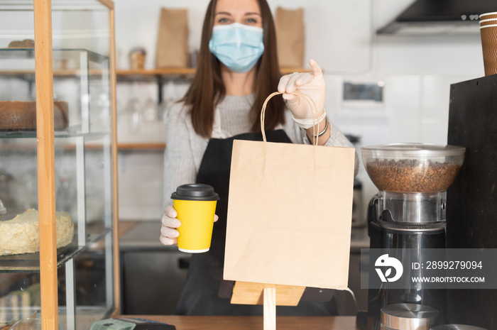 Package with a take away order and paper cup of coffee in hands of waitress, cafe owner, a woman in mask and gloves in out of focus. Protective measures due quarantine lockdown, pick up and go concept