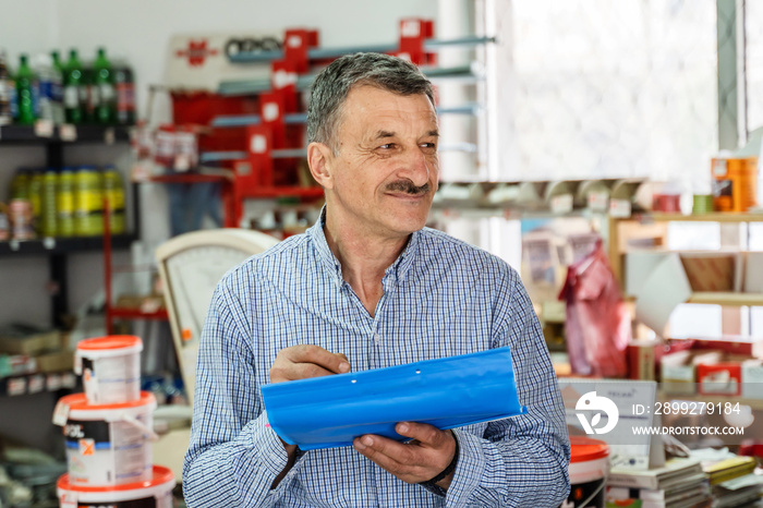 front view portrait of adult caucasian man senior entrepreneur at store holding document writing or making a list while looking to the side at hardware shop store real people small business concept