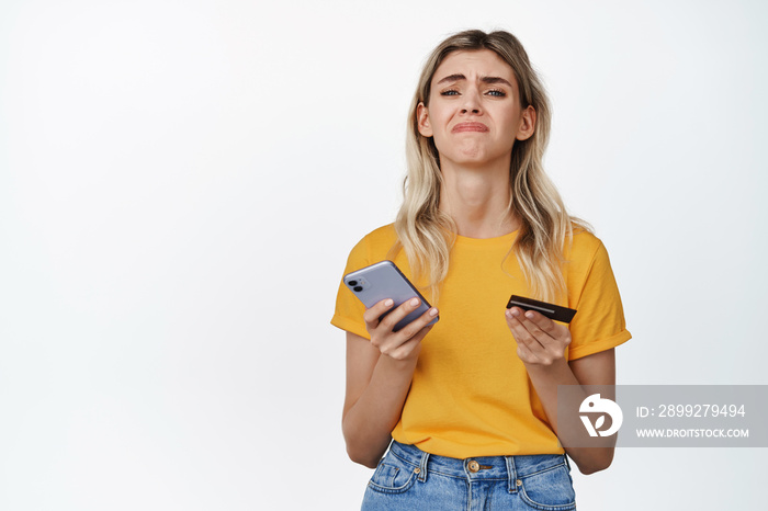 Upset girl holding smartphone and credit card, concept of woman lacking money, cant buy something, standing over white background