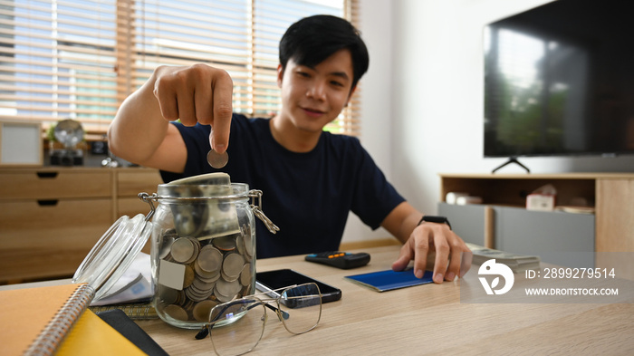 Smiling asian man putting coin into the glass jar, save money for future, retirement and financial planning concept