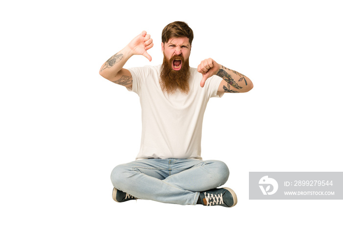 Young adult redhead with a long beard sitting on the floor isolated showing thumb down and expressing dislike.