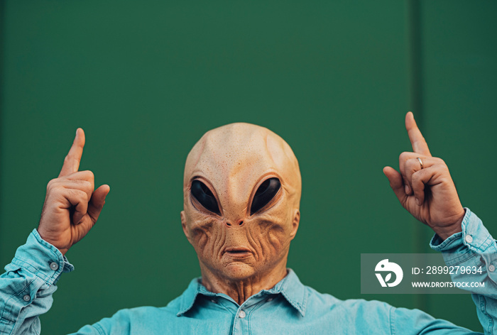 Portrait of ufo alien extraterrestrial mask with denim casual clothing like a human and green wall in background for copyspace text