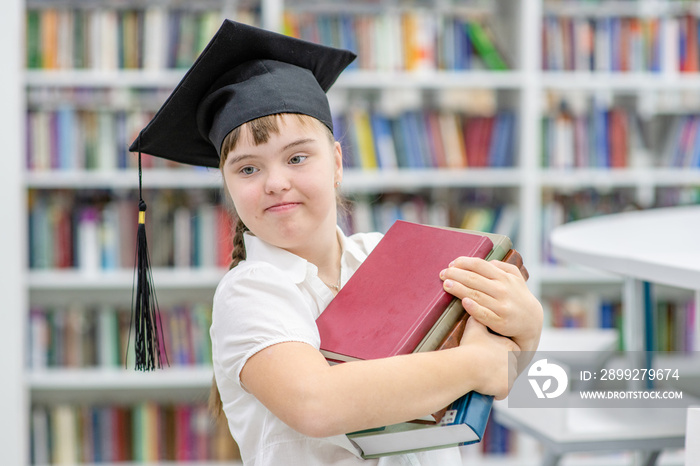 Happy girl with Down Syndrome wearing a graduation cap holds books in her hands at a library. Empty space for text