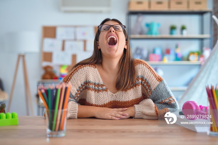 Young beautiful teacher woman wearing sweater and glasses sitting on desk at kindergarten angry and mad screaming frustrated and furious, shouting with anger. Rage and aggressive concept.