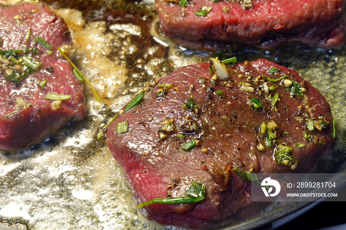 Venison steaks on frying pan. Deer meat sirloin is spiced with garlic, rosemary and thyme.