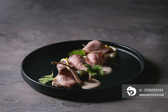 Vitello tonnato italian dish. Thin sliced veal with tuna sauce, capers and coriander served on black plate over dark texture background. Top view, space