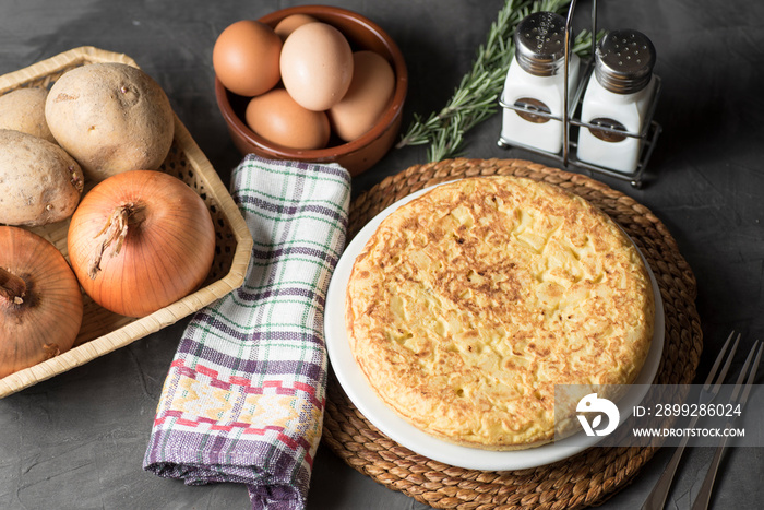 Spanish potato omelette, on a table, next to the necessary ingredients to be cooked and ready to be eaten