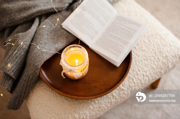 Scented burning candle with open paper book stay on wooden tray and knitted textile in chair top view close up. Cozy home atmosphere lifestyle.