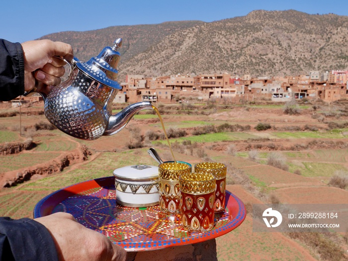 Man pouring mint tea into glasses on colorful tray, Ourika valley and High Atlas Mountains in the background. Morocco.