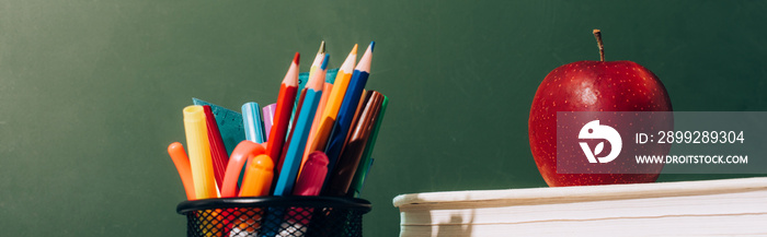 Website header of pen holder with color pencils and pens, and ripe apple on books near green chalkboard