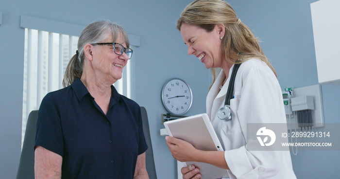 Senior patient laughing with her doctor in exam room. Female primary care physician talking to older Caucasian woman during regular check up