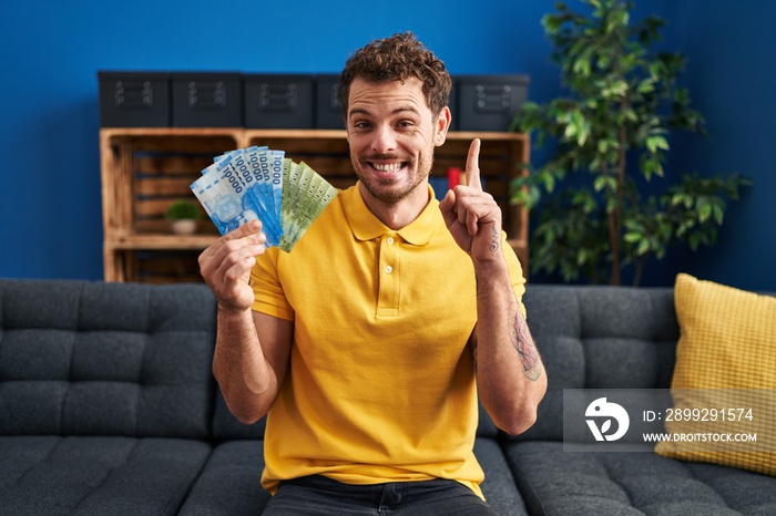 Young hispanic man holding chilean pesos surprised with an idea or question pointing finger with happy face, number one