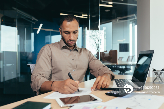 Serious thinking hispanic businessman working with documents and invoices inside office, man behind paper work, using laptop at work sitting at desk.